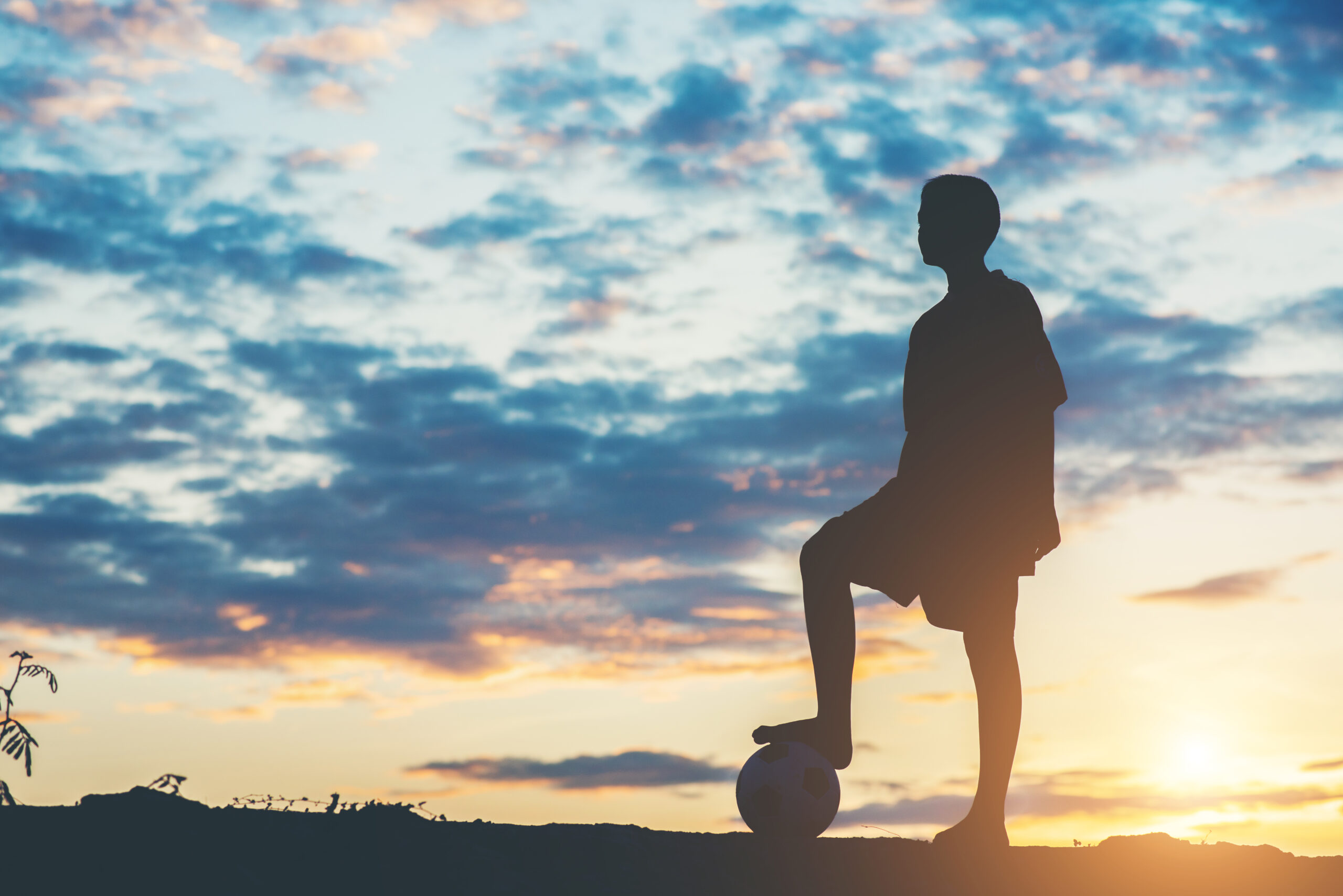 Young Filipino boy playing football barefoot on a dusty barangay pitch at sunset.