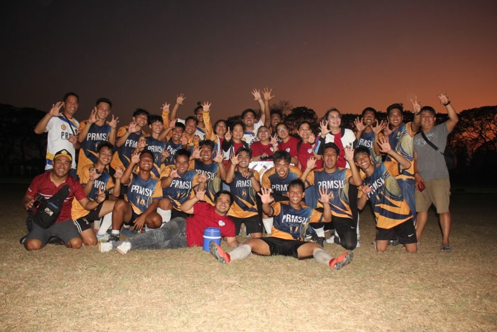 PRMSU football team celebrating together on the field at sunset, wearing blue and gold jerseys, showing school pride and unity.