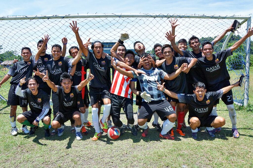 A joyful football team in black jerseys celebrates together in front of a goalpost, raising their hands and smiling after a match on a sunny day.