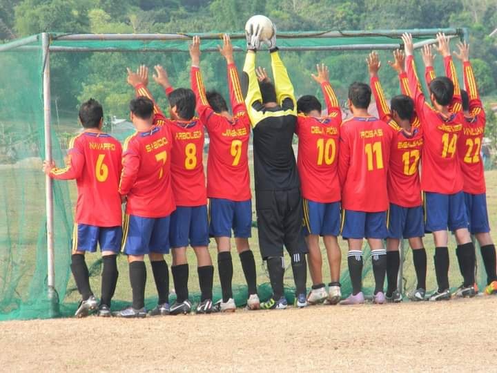 "Football team in red jerseys standing with raised hands at the goalpost during training, with the goalkeeper holding a ball overhead."