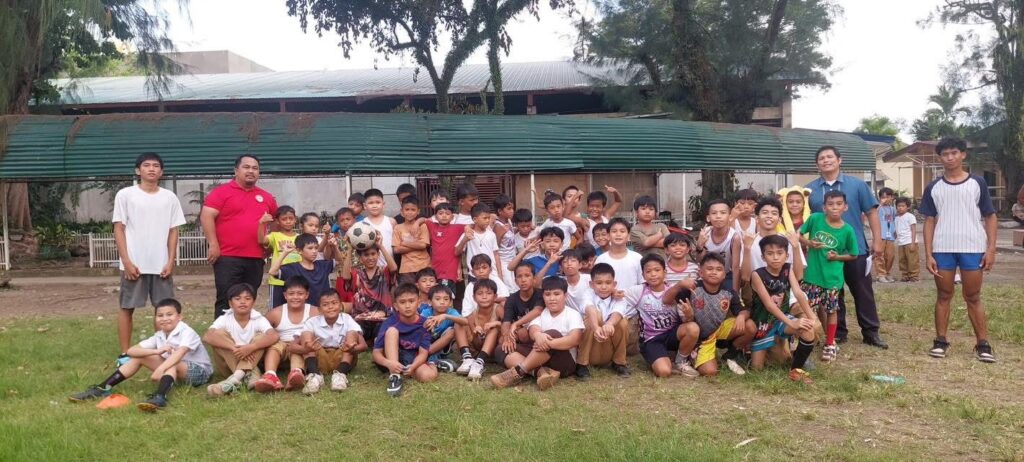 A large group of young boys in casual sportswear pose together on a grassy field along with three adult coaches. Some children are sitting in front while others stand behind them, smiling and holding a soccer ball. Trees and a simple building with a green roof are in the background.