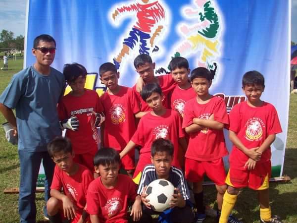 Young football players in red kits pose with their coach during a grassroots football event, with one child holding a football in front of a colorful tournament banner.