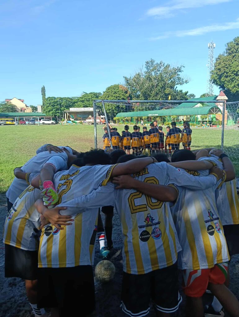 Bustos United Football Club players in white and gold jerseys form a tight huddle near the goalpost before a match on a grass field, showing unity in Philippine football.