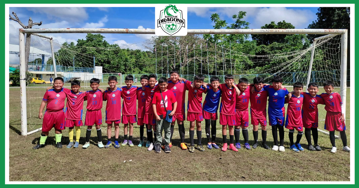 Green Dragons Football Club youth players in red and blue uniforms lined up in front of a goalpost during a sunny match day.