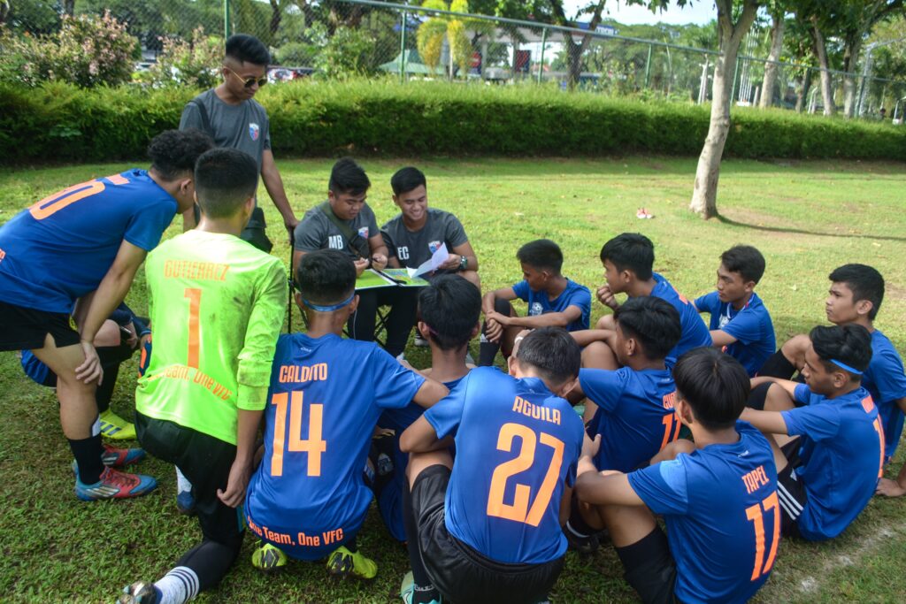 Villamor Football Club where Young football players in blue jerseys gathered near a large, textured tree trunk with natural hollow formations in an outdoor park.