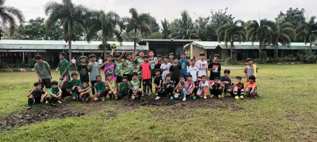 A large group of young football players from Villamor FC posing together on a grassy field after practice, wearing mixed football kits and sportswear in front of a school-like building lined with palm trees.